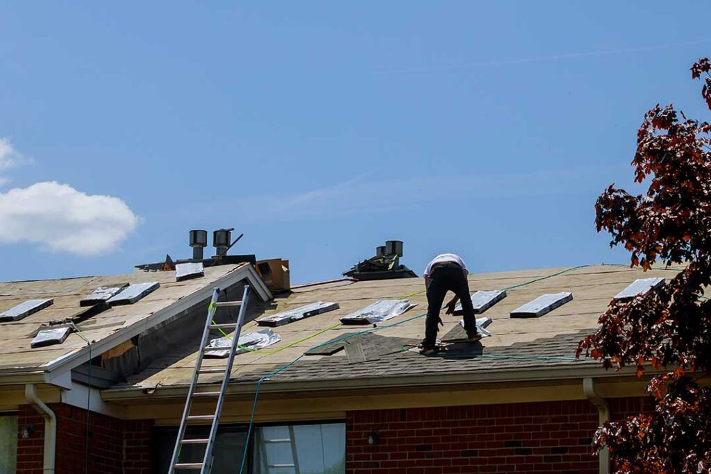 A person cleaning debris from an asphalt shingle roof using a soft brush and gloves.