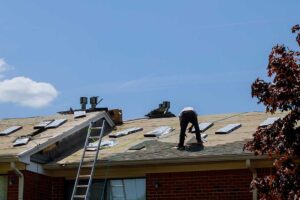 A person cleaning debris from an asphalt shingle roof using a soft brush and gloves.
